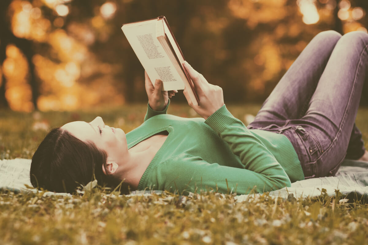 woman lying on a rug outdoors reading a book.