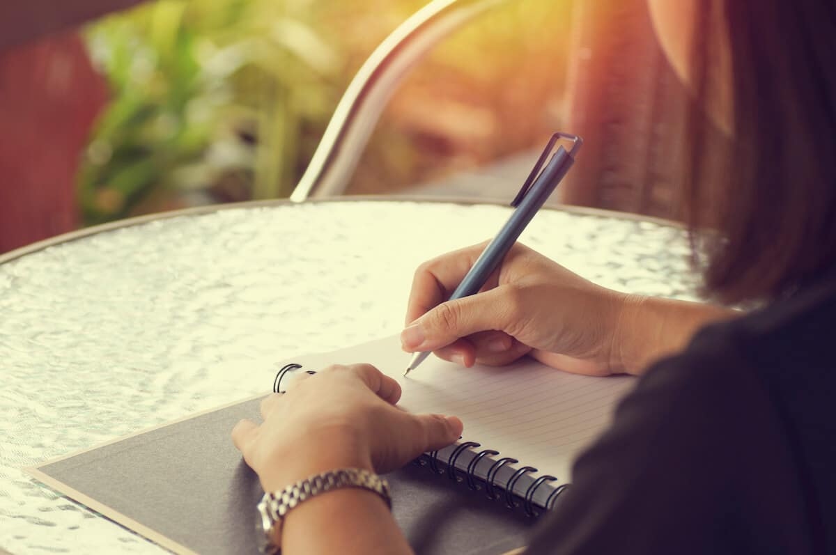 woman sitting at a table in the sun writing in a journal.