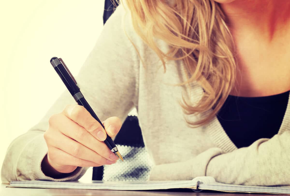 woman sitting at a table in a white jacket writing in a note book.