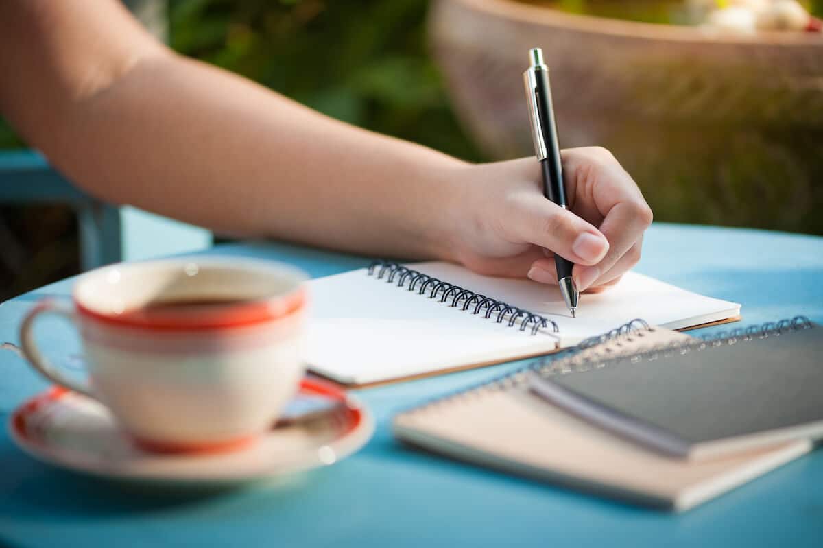 woman at a blue table with a cup of tea writing in a journal.