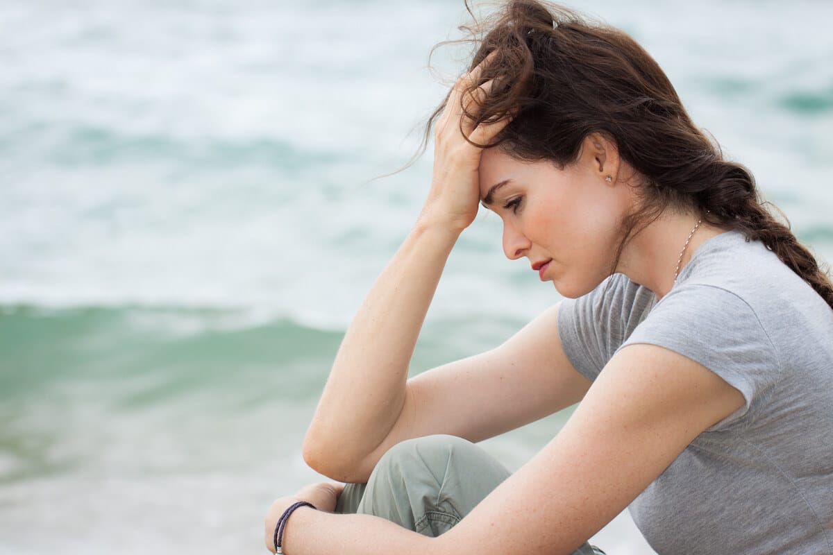 upset woman holding her head while sitting near the ocean.