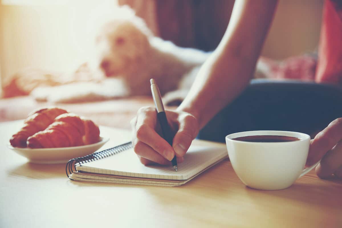 woman sitting on the floor writing in a notebook with a dog in the background and a plate with a croisant.