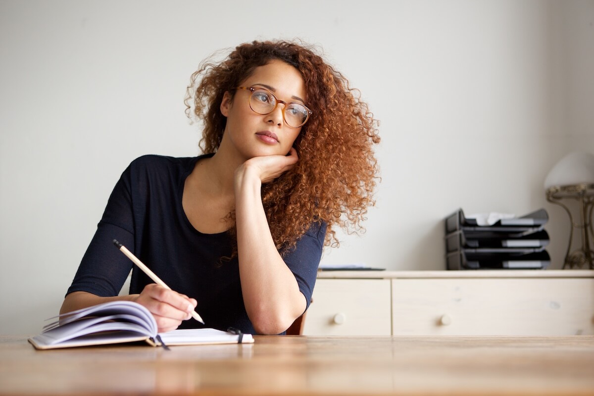 woman sitting at desk, thinking and writing in journal.