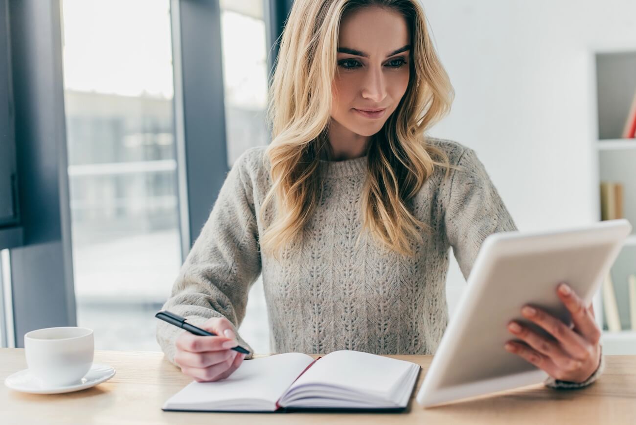 woman looking at tablet while writing in her journal.