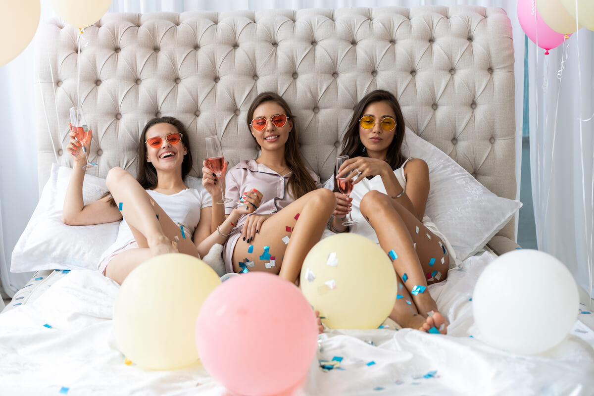 three women sitting in a bed with balloons and drinking champagne.